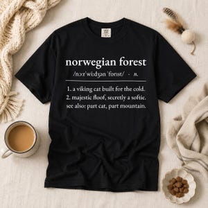 May include: A black short-sleeved t-shirt featuring white text that reads: norwegian forest /nɔːr'wiːdʒən 'fɔrɪst/ · n. 1. a viking cat built for the cold. 2. majestic floof, secretly a softie. see also: part cat, part mountain.