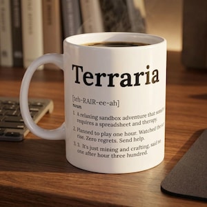 Può includere: Tazza in ceramica bianca con la scritta "Terraria" in nero. La tazza contiene un liquido scuro e una definizione del gioco. La tazza è su una superficie di legno, con una tastiera e libri sullo sfondo. La definizione è in inglese.