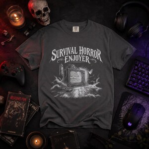 Könnte beinhalten: Ein anthrazitfarbenes Kurzarm-T-Shirt mit einem weißen Grafikdruck eines alten Fernsehers auf einem Holzboden und dem darüber gewölbten Text SURVIVAL HORROR ENJOYER. Das Design zeigt wirbelnden Nebel und einen kleinen Vogel am Bildschirm.