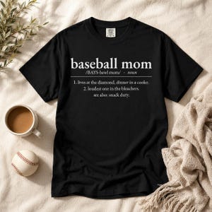 以下が含まれることがあります： 白い文字がプリントされた黒の半袖Tシャツ。文字の内容は「baseball mom /BAYS-bawl mom/ noun 1. lives at the diamond, dinner in a cooler. 2. loudest one in the bleachers. see also: snack duty.」です。クルーネックで、襟元にタグが付いています。