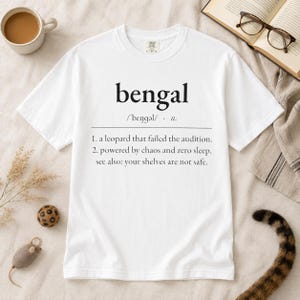 May include: A white short-sleeve t-shirt featuring black text that reads: bengal /'bɛŋɡəl/ n. 1. a leopard that failed the audition. 2. powered by chaos and zero sleep. see also: your shelves are not safe. The shirt has a crew neckline and a standard fit.
