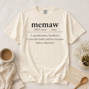 Könnte beinhalten: Ein cremefarbenes Kurzarm-T-Shirt mit schwarzem Text, der das Wort memaw als eine Großmutter aus den Südstaaten definiert, die die Familie leitet und jeden glauben lässt, es sei anders. Das T-Shirt hat einen Rundhalsausschnitt.