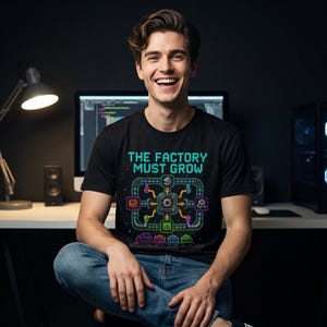 Op de afbeelding: Een zwart t-shirt met korte mouwen voorzien van een kleurrijke pixel art afbeelding van een fabrieksassemblagelijn met de tekst "THE FACTORY MUST GROW" in een retro digitaal lettertype, inclusief iconen van tandwielen, buizen en een treintje.