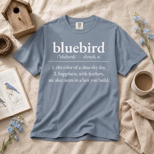 May include: A slate blue short-sleeve t-shirt featuring white text that reads: bluebird /'blubərd/ - thrush, n. 1. the color of a clear-sky day. 2. happiness, with feathers. see also: nests in a box you build. The shirt has a crew neckline.