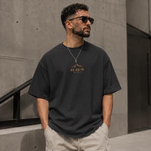 May include: A charcoal grey short-sleeve t-shirt featuring a small graphic on the chest. The graphic depicts a mountain range in orange above the text "PS 46:10" and "BE STILL" in a matching orange font. The shirt has a crew neckline and a loose fit.