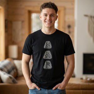 以下が含まれることがあります： 黒の半袖Tシャツで、中央に3つの灰色の石のような形が縦に並んだグラフィックがプリントされています。それぞれの石には、濃い色の記号が刻まれています。Tシャツはクルーネックで、青いデニムジーンズと組み合わされています。