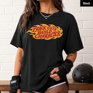 Könnte beinhalten: Schwarzes T-Shirt mit dem Text "TRACK CUTS BUILD CHARACTER" in einem flammenartigen Design. Der Text ist in Gelb und Rot gehalten. Das Shirt wird mit schwarzen Shorts und Schutzkleidung getragen.
