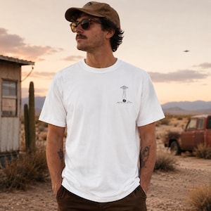 Könnte beinhalten: Weißes T-Shirt mit kurzen Ärmeln und einer kleinen schwarz-weißen Grafik eines UFOs. Das T-Shirt wird von einer Person in einer Wüstenlandschaft getragen. Die Person trägt eine braune Kappe und Sonnenbrille.