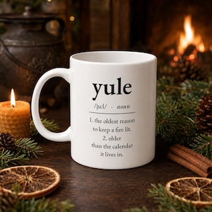 May include: A white ceramic mug featuring black text that defines the word yule as the oldest reason to keep a fire lit and older than the calendar it lives in. The mug has a standard handle and a smooth finish with clear, legible typography.