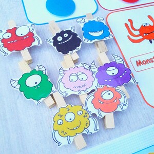 May include: A set of colorful monster illustrations with different facial expressions. Each monster is attached to a wooden clothespin. The monsters are red, black, blue, green, pink, purple, orange, and yellow. The text on the cards reads "Monstre bleu" and "Monstre rouge".
