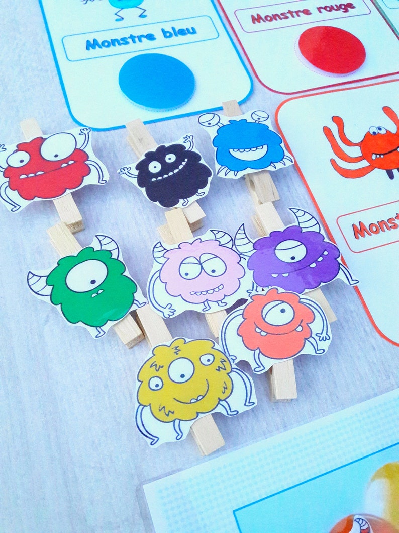 May include: A set of colorful monster illustrations with different facial expressions. Each monster is attached to a wooden clothespin. The monsters are red, black, blue, green, pink, purple, orange, and yellow. The text on the cards reads "Monstre bleu" and "Monstre rouge".