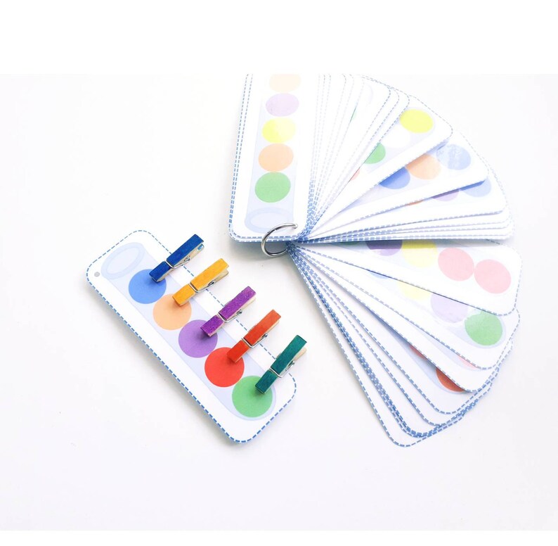 Puede incluir: Un juego de tarjetas de coincidencia de colores con una variedad de colores, incluyendo rojo, naranja, amarillo, verde, azul, morado y rosa. Las tarjetas est&aacute;n dispuestas en forma de abanico y tienen un borde azul punteado. Tambi&eacute;n hay seis pinzas para la ropa de colores.