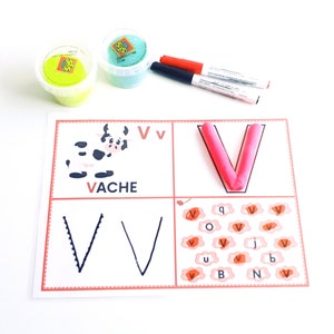 May include: A white card with a red border and the letter V printed on it. The letter V is also formed with pink playdough. There are several other Vs printed on the card, as well as the word VACHE.