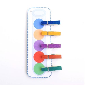 Puede incluir: Una actividad de aprendizaje colorida con una tarjeta blanca que presenta un tubo de ensayo con cinco c&iacute;rculos de colores. Los c&iacute;rculos son azul, naranja, morado, rojo y verde. Hay cinco pinzas de madera, cada una pintada de un color diferente para que coincida con los c&iacute;rculos. Las pinzas est&aacute;n sujetas a la tarjeta.