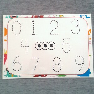 May include: A white sheet of paper with a red border and a dotted line outline of the numbers 0 through 9. There are three black circles in the center of the sheet.
