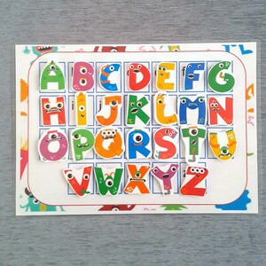 May include: A colorful alphabet chart with each letter represented by a cartoon monster. The letters are arranged in a grid with a white background and a colorful border.
