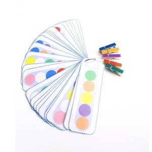 Puede incluir: Un juego de tarjetas de coincidencia de colores con una variedad de colores, incluyendo rojo, naranja, amarillo, verde, azul, morado y rosa. Las tarjetas est&aacute;n dispuestas en forma de abanico y se mantienen unidas por un anillo de metal. Tambi&eacute;n hay un juego de pinzas para la ropa de colores.