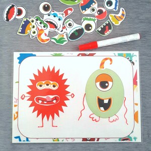 May include: A white card with two cartoon monsters on it. The monster on the left is red with three eyes and a tongue sticking out. The monster on the right is green with one eye and a toothy grin. There are colorful monster stickers scattered around the card.