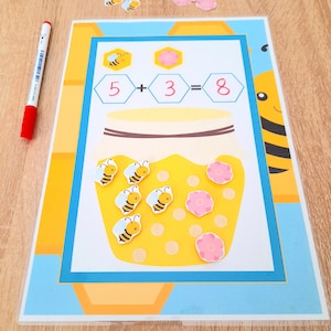 Puede incluir: Un juego de aprendizaje amarillo y azul con un tarro de miel y abejas. El juego incluye una ecuación matemática: 5 + 3 = 8. Hay pegatinas de abejas y flores para colocar en el tarro de miel.