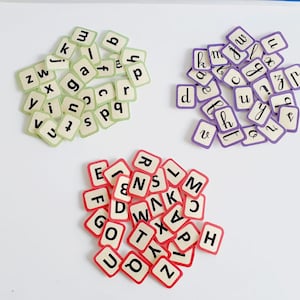 May include: Three piles of letter tiles, each in a different colour. The first pile is green, the second is purple, and the third is red. The tiles are all white with black letters.