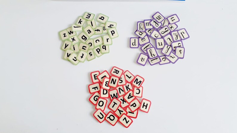 May include: Three piles of letter tiles, each in a different colour. The first pile is green, the second is purple, and the third is red. The tiles are all white with black letters.