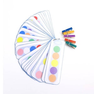 Puede incluir: Un juego de tarjetas de coincidencia de colores con una variedad de colores, incluyendo rojo, naranja, amarillo, verde, azul, morado y rosa. Las tarjetas est&aacute;n dispuestas en forma de abanico y tienen un c&iacute;rculo blanco en el centro de cada tarjeta. Tambi&eacute;n hay cinco pinzas para la ropa de colores.