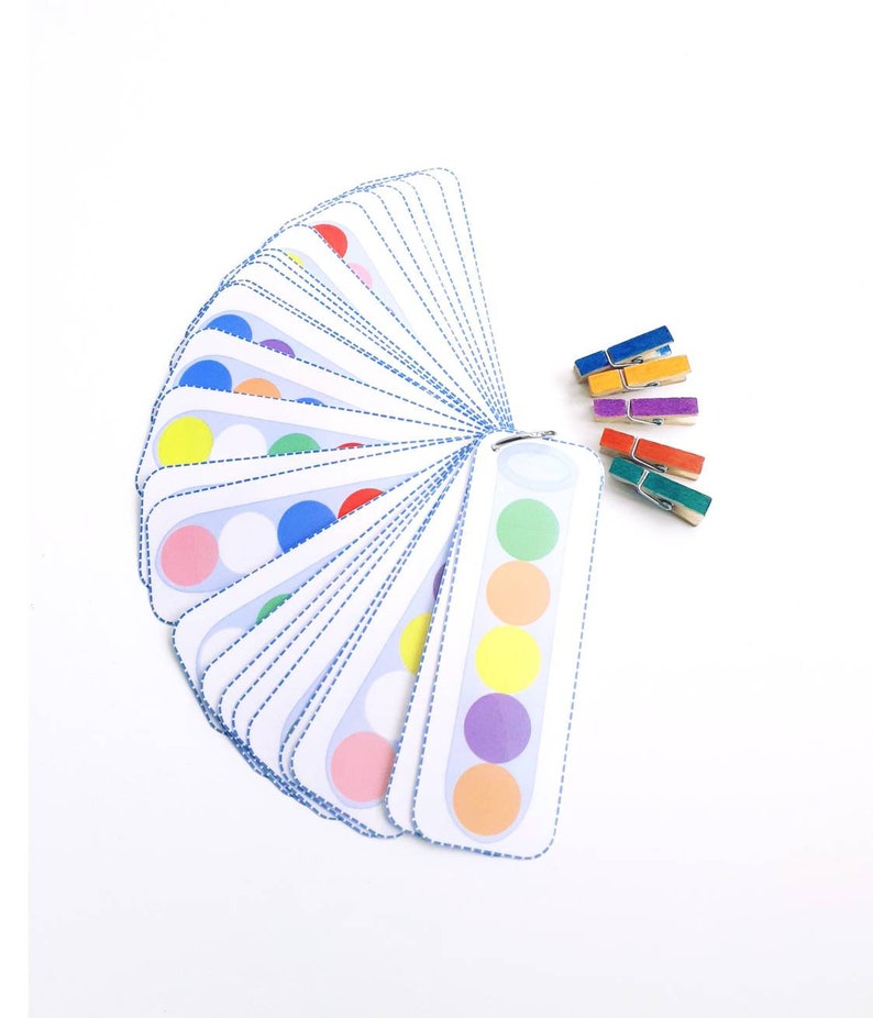 Puede incluir: Un juego de tarjetas de coincidencia de colores con una variedad de colores, incluyendo rojo, naranja, amarillo, verde, azul, morado y rosa. Las tarjetas est&aacute;n dispuestas en forma de abanico y tienen un c&iacute;rculo blanco en el centro de cada tarjeta. Tambi&eacute;n hay cinco pinzas para la ropa de colores.