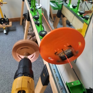 May include: An orange and brown spooling machine with a yellow drill. The machine has a wooden frame and green components. A bright orange line is being spooled onto a black rod. The machine is set up in a workshop.