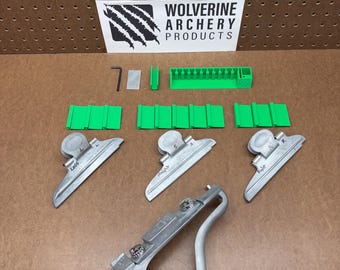 May include: An archery fletching jig kit including a silver metal base, three silver metal clamps labeled Left, Straight, and Right, three green plastic clamp bases, a green plastic rack, a small metal hex key, and a rectangular metal plate.
