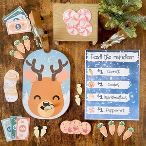 May include: A game for kids to feed a reindeer with different treats. The game includes a reindeer head with a slot for the treats, a set of carrots, cookies, marshmallows, and peppermint candies, and a price tag for each treat. The game also includes a set of dollar bills and a set of snowflakes.