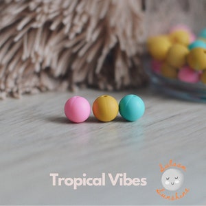 May include: Three silicone beads in pink, yellow, and blue. The beads are round and smooth. They are on a light wood surface.