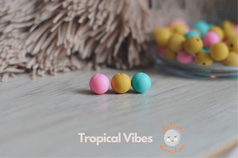 May include: Three silicone beads in pink, yellow, and blue. The beads are round and smooth. They are on a light wood surface.