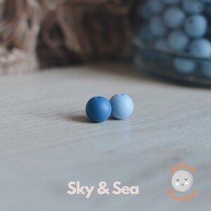 May include: Two silicone beads, one dark blue and one light blue, on a white surface. The beads are round and smooth. The text "Sky & Sea" is visible below the beads.
