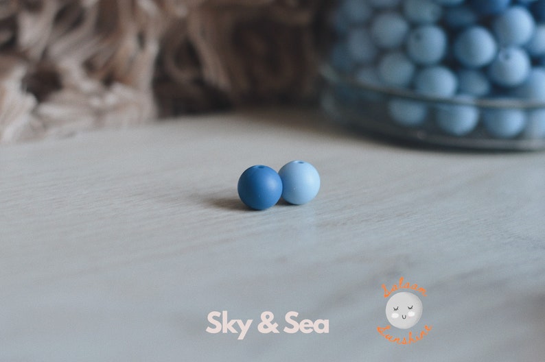 May include: Two silicone beads, one dark blue and one light blue, on a white surface. The beads are round and smooth. The text "Sky & Sea" is visible below the beads.