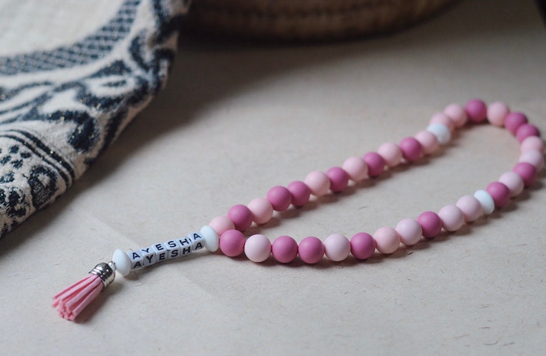 May include: A pink and white beaded necklace with a tassel and a white bead with the name "AYESHA" spelled out on it.