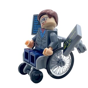 May include: A Lego minifigure of a person in a wheelchair, wearing glasses, a gray suit, and a blue shirt. The figure has brown hair and is holding two black screens. The wheelchair has black wheels and a gray frame.