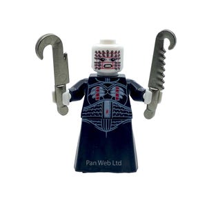 May include: A Lego minifigure of a character with a white head featuring a pattern of red dots and a grimacing mouth. The figure wears a black outfit with a detailed design and holds two silver, curved blades. The text "Pan Web Ltd" is visible.
