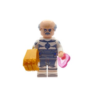 May include: A Lego minifigure of a character with a light skin tone, gray hair, and a gray mustache. The figure wears a white and gray suit and holds a gold gauntlet and a pink heart-shaped object. The figure is standing on a white surface.