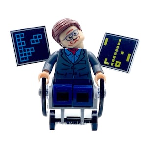 May include: A Lego minifigure of a man in a wheelchair, wearing glasses, a suit, and a red tie. Two black and blue game screens are behind the figure. The figure has brown hair and a serious expression.