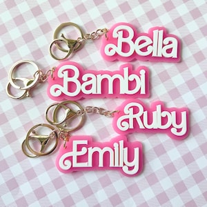 Puede incluir: Llaveros rosas de acrílico con letras blancas que deletrean los nombres Bella, Bambi, Ruby y Emily. Cada llavero tiene un cierre y una cadena dorados. Los llaveros se muestran sobre un fondo a cuadros rosa y blanco.