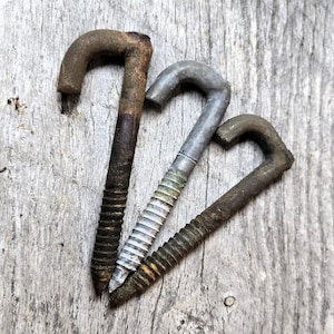 May include: Three vintage metal hook bolts with threaded shafts. The hooks are curved and show signs of age, with varying degrees of rust and wear. The bolts are arranged on a weathered wooden surface.