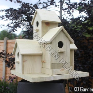 May include: A multi-compartment wooden birdhouse with a natural finish. The birdhouse features multiple levels, round entry holes, and a small perch below each hole. The design includes a flat base and a pitched roof. The birdhouse is outdoors.