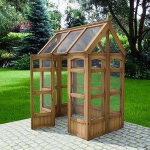 May include: A wooden greenhouse with a peaked roof and glass panels. The structure has a door open to the left, revealing the interior. The greenhouse is set on a stone patio, surrounded by green grass and trees.