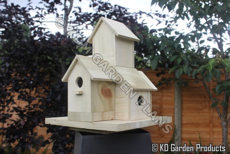 May include: A wooden birdhouse with a multi-level design. The birdhouse has a rectangular base and a series of gabled roofs. The birdhouse has a natural wood color and features circular entry holes. The text "KD Garden Plans" is visible.