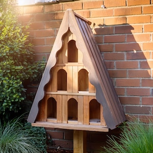 Op de afbeelding: Een houten vogelhuisje met een driehoekig dak en meerdere nestvakken. Het vogelhuisje is gemonteerd op een houten paal en heeft een donkerbruine vlek.