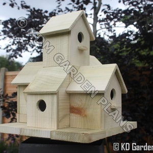 May include: A multi-compartment wooden birdhouse with multiple gabled roofs and circular entry holes. The birdhouse is made of light-colored wood and is designed to attract birds. The text "KD Garden Plans" is visible on the side.