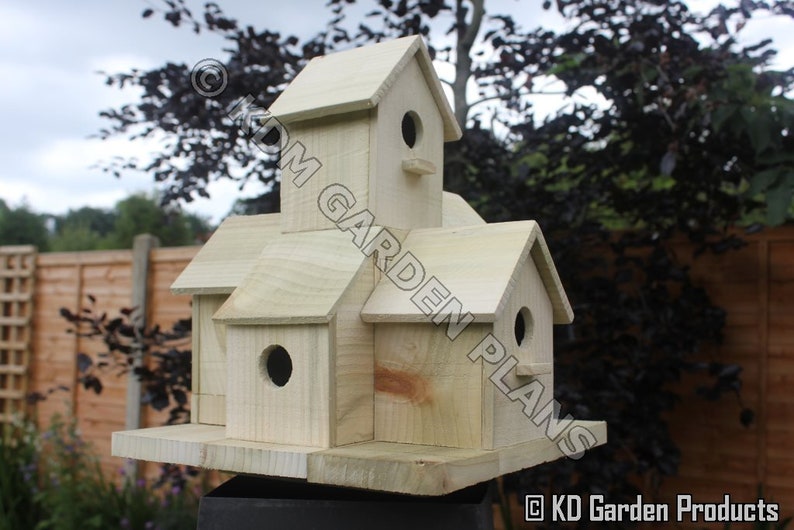 May include: A multi-compartment wooden birdhouse with multiple gabled roofs and circular entry holes. The birdhouse is made of light-colored wood and is designed to attract birds. The text "KD Garden Plans" is visible on the side.