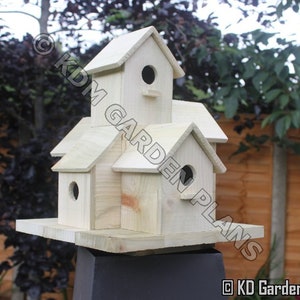 May include: A multi-level wooden birdhouse with multiple compartments and circular entry holes. The birdhouse is constructed from light-colored wood and features a pitched roof design. The birdhouse is mounted on a black stand, with a natural outdoor background.
