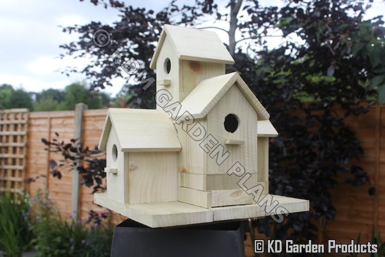 May include: A multi-compartment wooden birdhouse with a natural finish. The birdhouse has multiple levels and circular entry holes. The design includes a small perch below each hole. The birdhouse is mounted on a black stand and set against a backdrop of greenery.