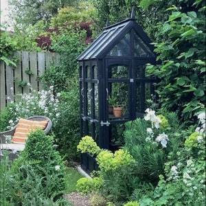 May include: A black wooden greenhouse with glass panels sits in a lush garden setting. The greenhouse is surrounded by green plants and white flowers. A small path leads to the greenhouse.